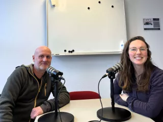 Twee personen in een lokaal met podcastmicrofoons en een white board aan de muur op de achtergrond